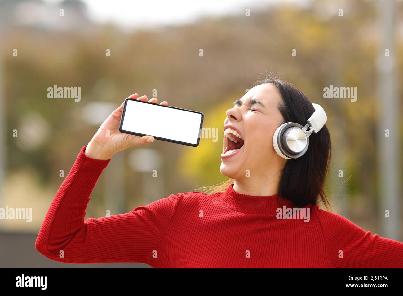 Happy woman in red wearing headphones singing in the street with blank smart phone screen Stock Photo