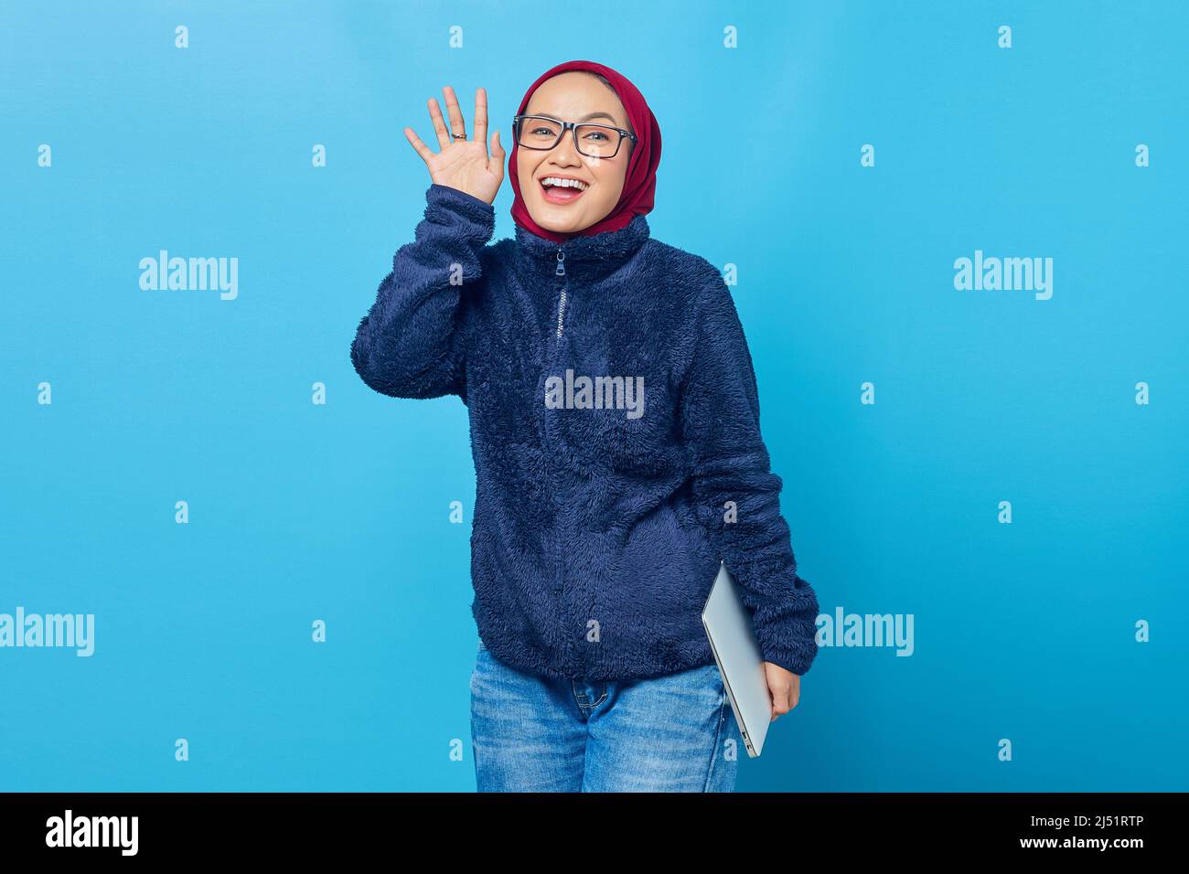 Smiling beautiful student holding laptop while waving to camera on blue background Stock Photo