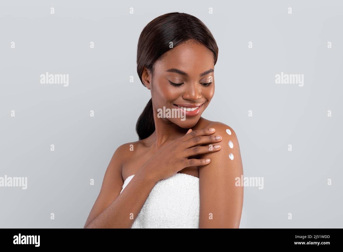 Cheerful pretty young black female in towel after shower applies cream to her shoulder, isolated on gray background Stock Photo