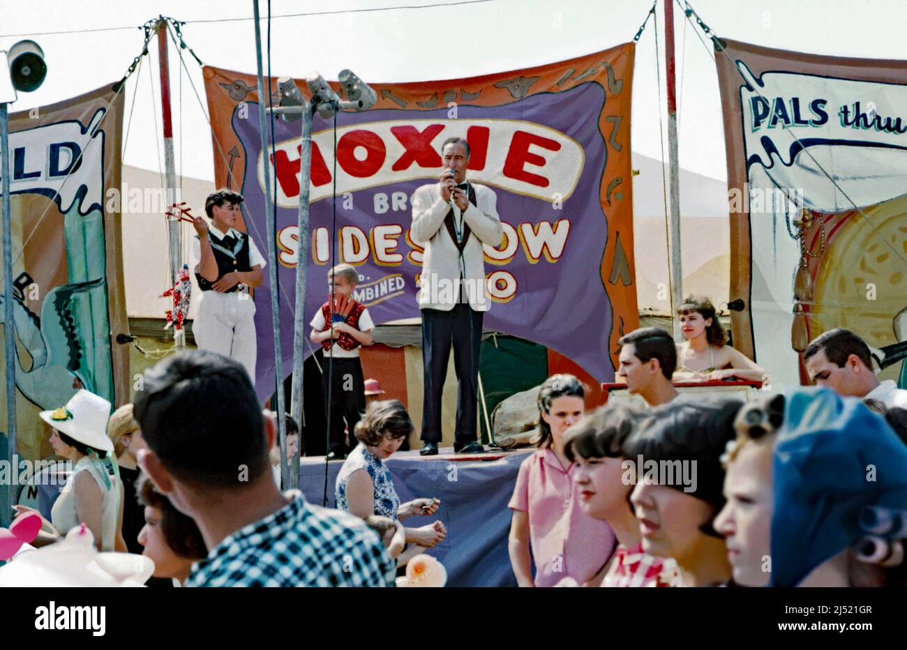 An announcer or barker with a microphone addressing the passing crowds outside the Hoxie Brothers Sideshow and Combined Zoo at its travelling circus in the USA in 1965. Alongside him is a boy holding knives and a young man with two clown puppets. Behind are colourful hand-painted banners advertising the attractions. This image is from an old American amateur (badly damaged) colour transparency. It has been heavily retouched and will look its best used at small sizes – a vintage 1960s photograph. Stock Photo