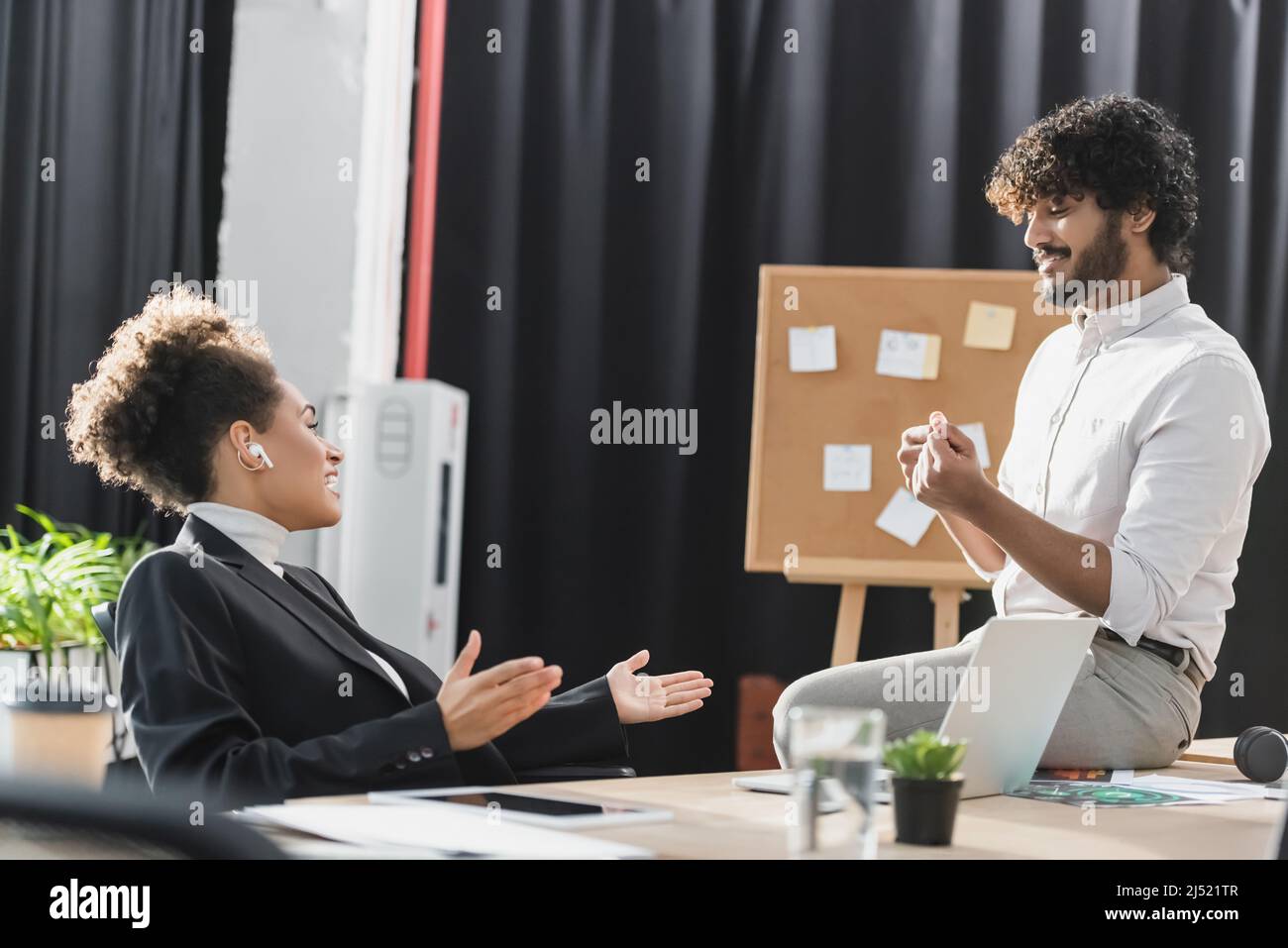 Side view of cheerful african american businesswoman in earphone talking to indian colleague near gadgets in office Stock Photo