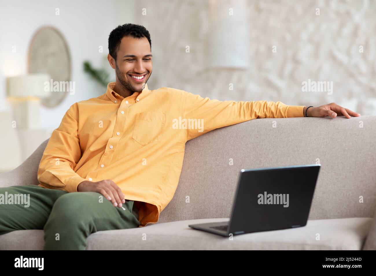 Smiling young man watching video on computer, sitting on couch Stock Photo