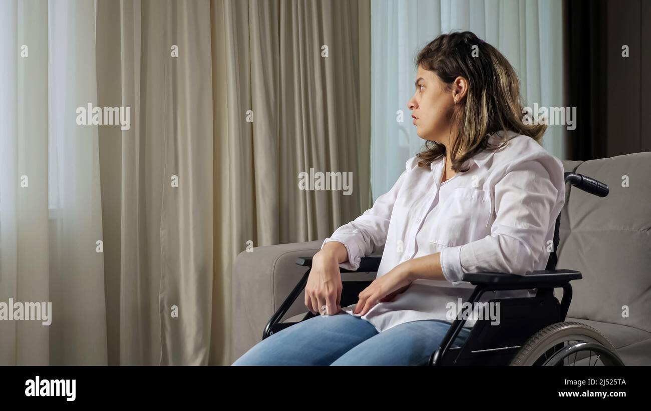 Sad disabled woman in wheelchair looks out of window at home Stock Photo
