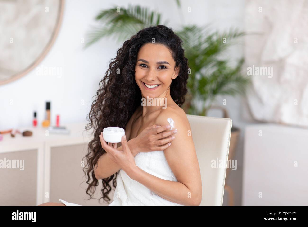 Smiling young caucasian brunette woman with long curly hair in towel applies cream on shoulder in bedroom interior Stock Photo