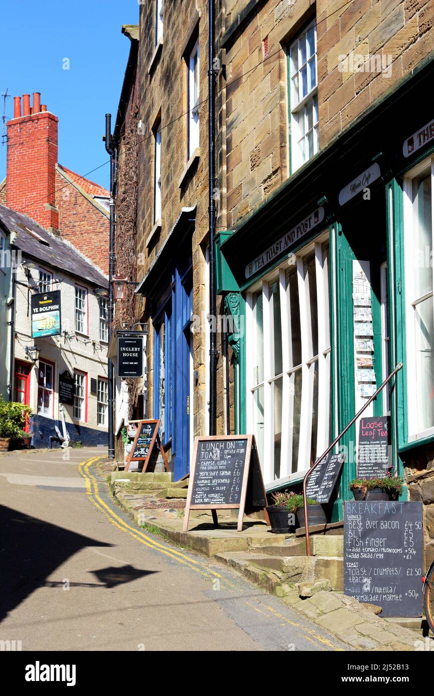 A charming street winding through the village of Robin's Hood Bay, including the Tea Toast and Post cafe, Ye Dolphin pub, shops and holiday lets Stock Photo