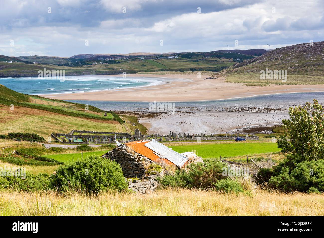 An old stone building with a tin roof overlooks the large, deserted, sandy beach and brilliant turquoise sea at Torrisdale Bay, on a sunny day. Stock Photo
