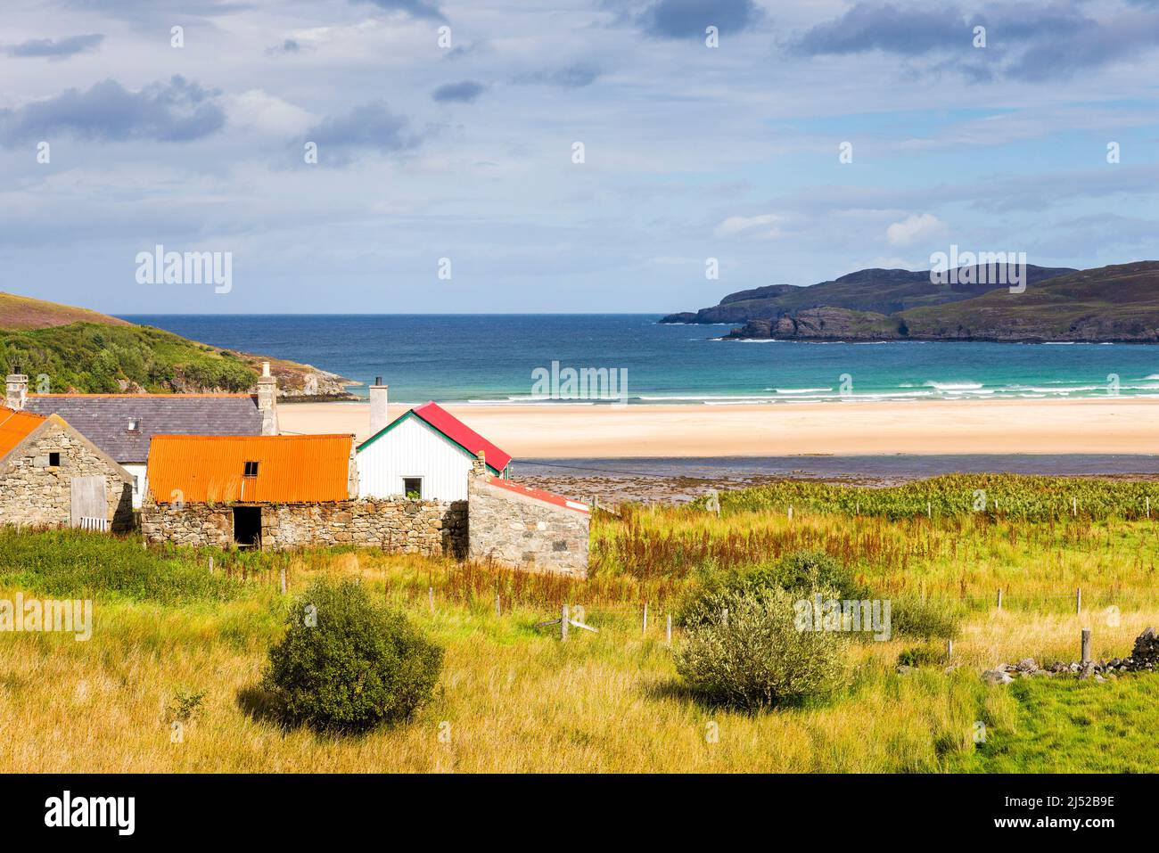 A small cluster of old farm buildings overlook a large, deserted, sandy beach and brilliant turquoise sea at Torrisdale Bay, on a sunny day. Stock Photo