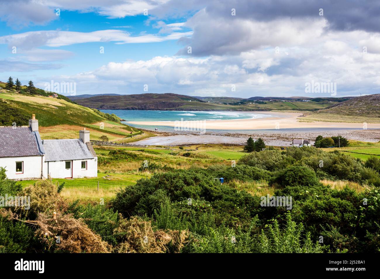 A white cottage on a grassy hill overlooks a large, deserted, sandy beach at Torrisdale Bay, on a sunny day. Stock Photo