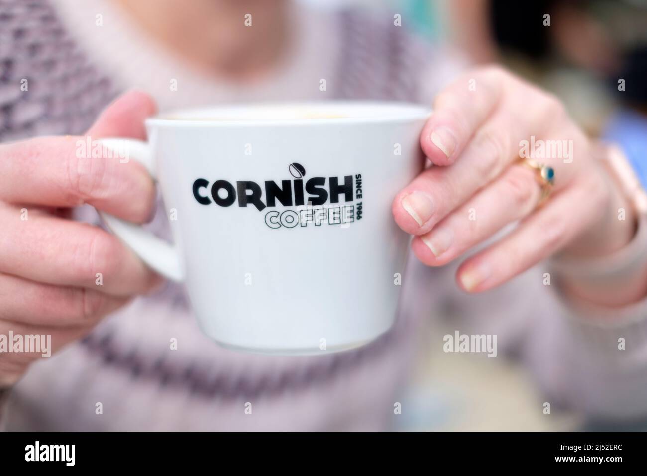 A woman holds a freshly made cup of Cornish coffee outside a cafe in Newquay, Cornwall, UK. The coffee cup has a Cornish Coffee Ltd company logo Stock Photo