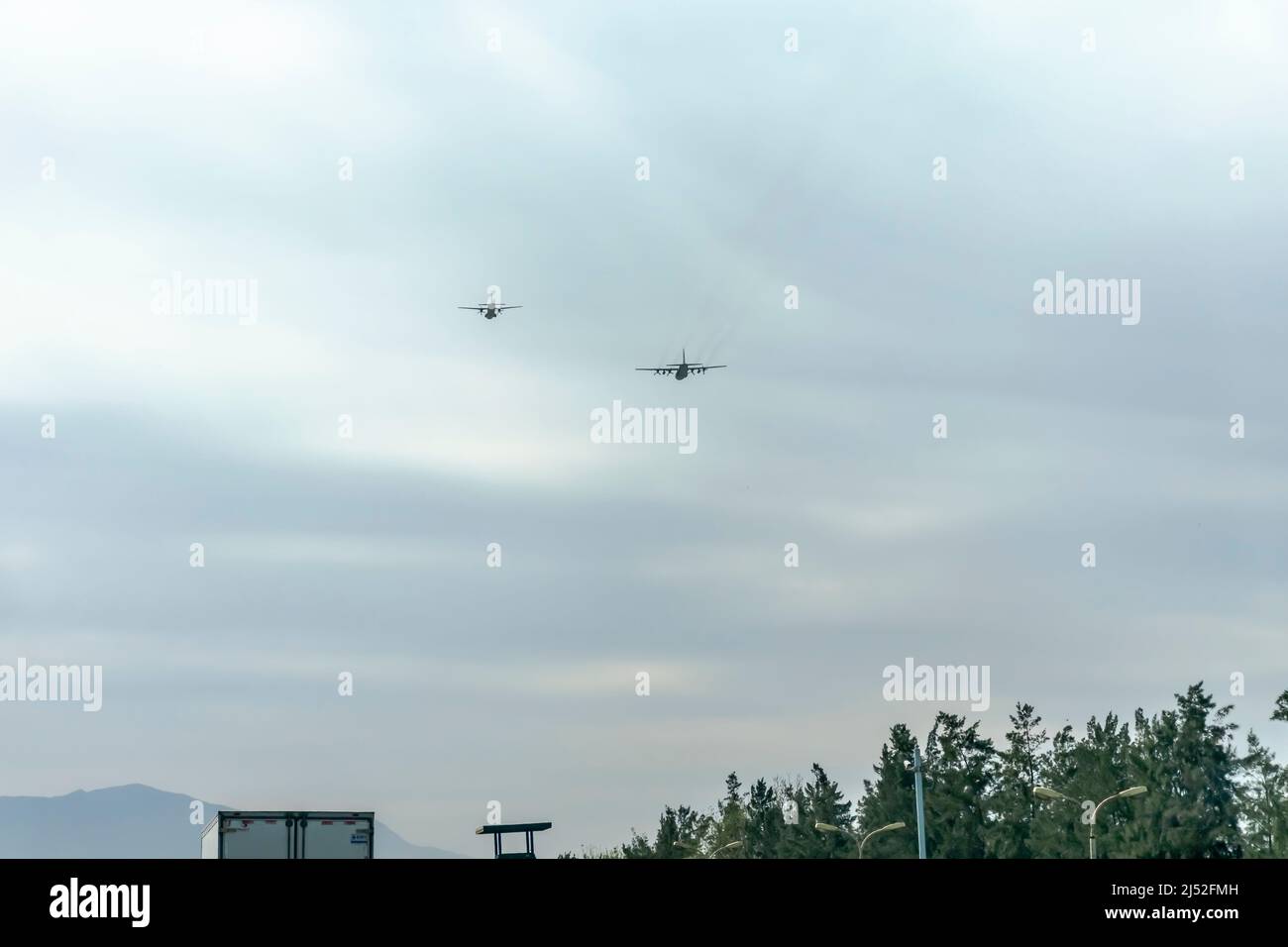 Two airplanes flying above Blida highway on Boufarik military airport. Overcast covered cloudy sky. Par of a truck, trees, and the mountain summit. Stock Photo