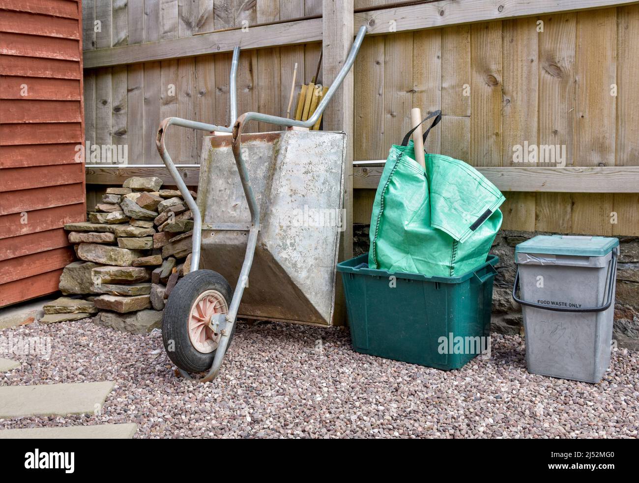 Garden corner with gardening items including wheelbarrow, large spade, large fork, Stone pile, gloves and wellies against wooden fence and stone wall Stock Photo