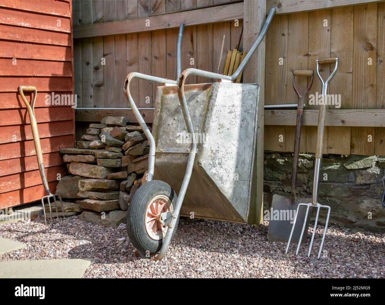 Garden corner with gardening items including wheelbarrow, large spade, large fork, Stone pile, gloves and wellies against wooden fence and stone wall Stock Photo