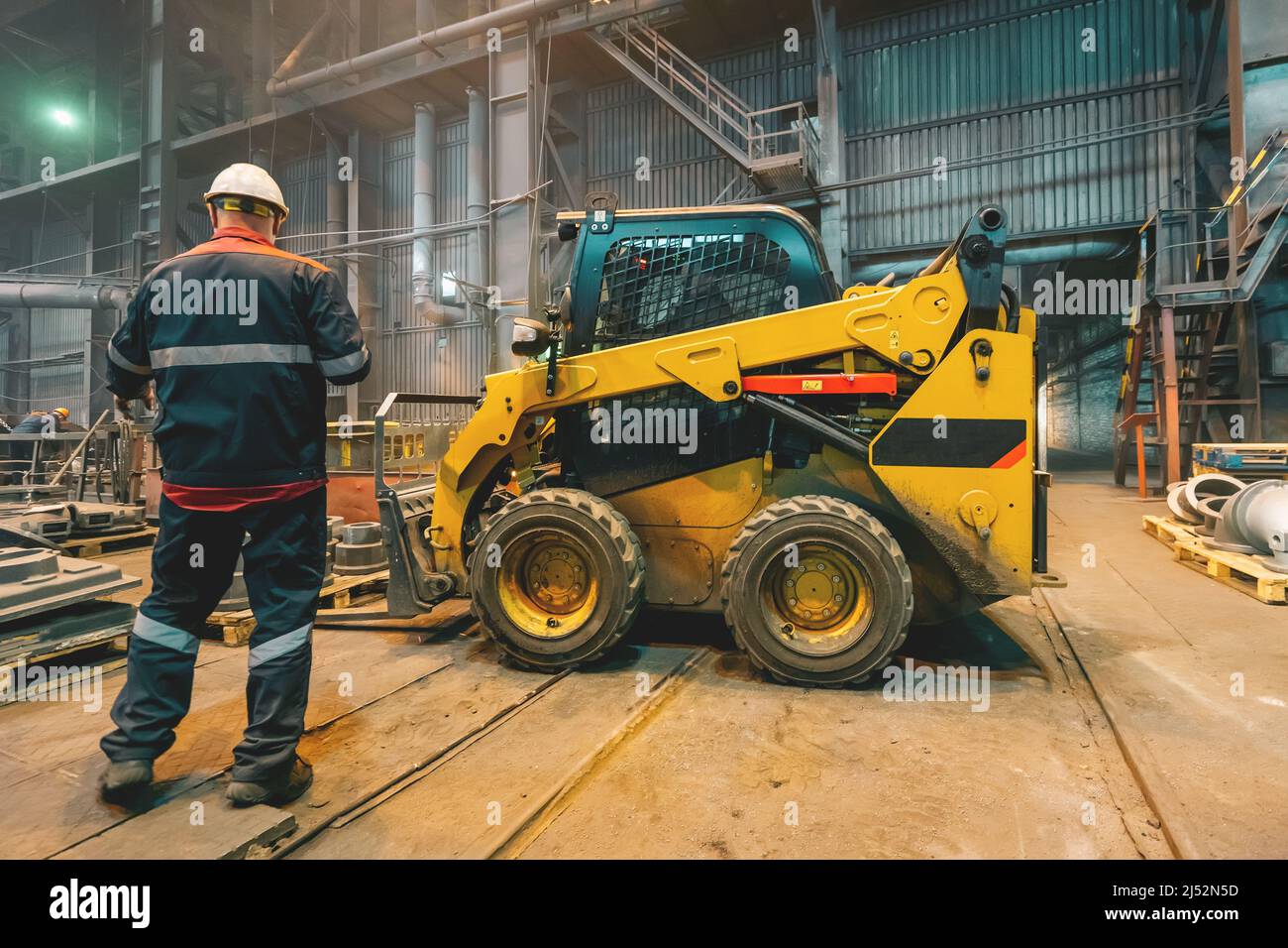 Workshop of metallurgical plant with small loading tractor or mower and unrecognizable engineer. Stock Photo