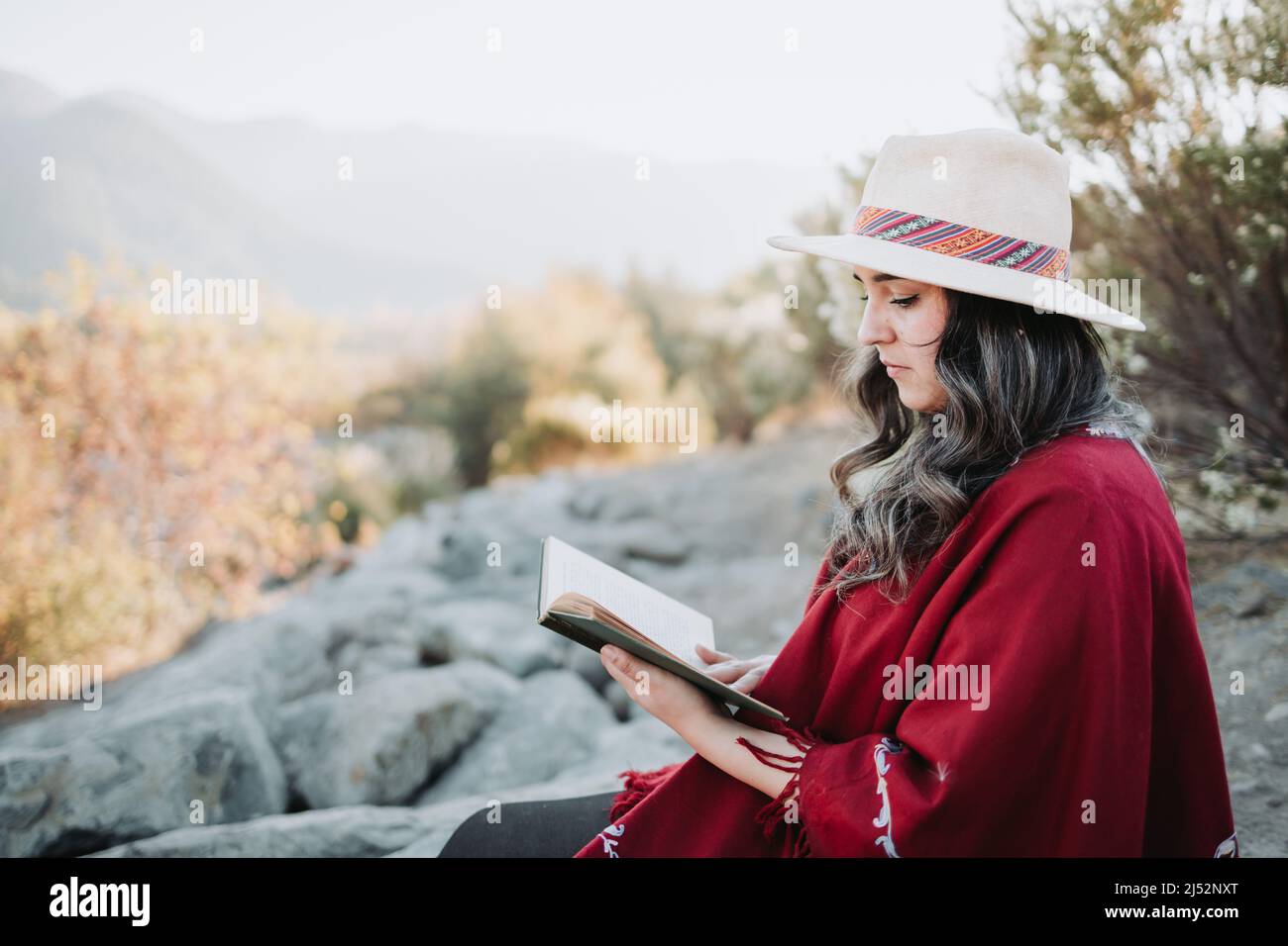 Young latin woman using a hat and a red poncho, sitting outdoor and reading a book. Copy space. Stock Photo