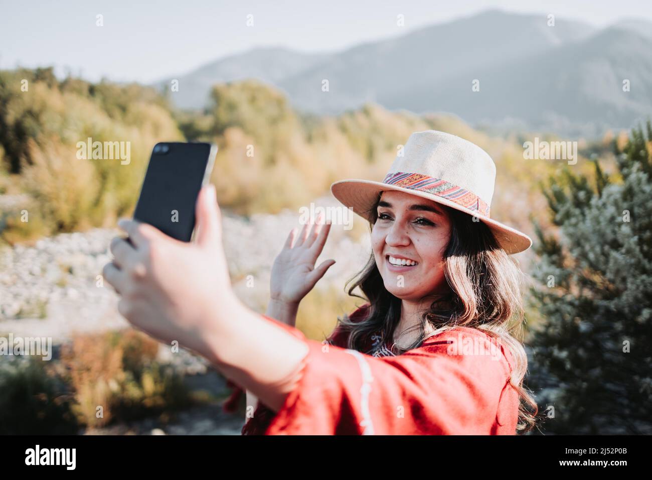 Beautiful and smiling young traveler woman making a video call from her mobile in a natural space. Stock Photo