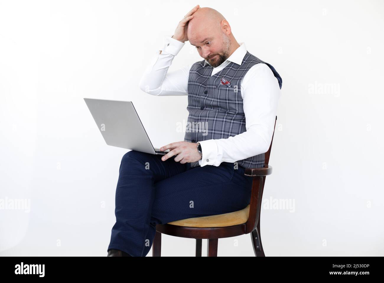 Portrait of bewildered middle-aged man in grey checkered vest, blue jeans, white shirt, sitting on chair holding laptop. Stock Photo