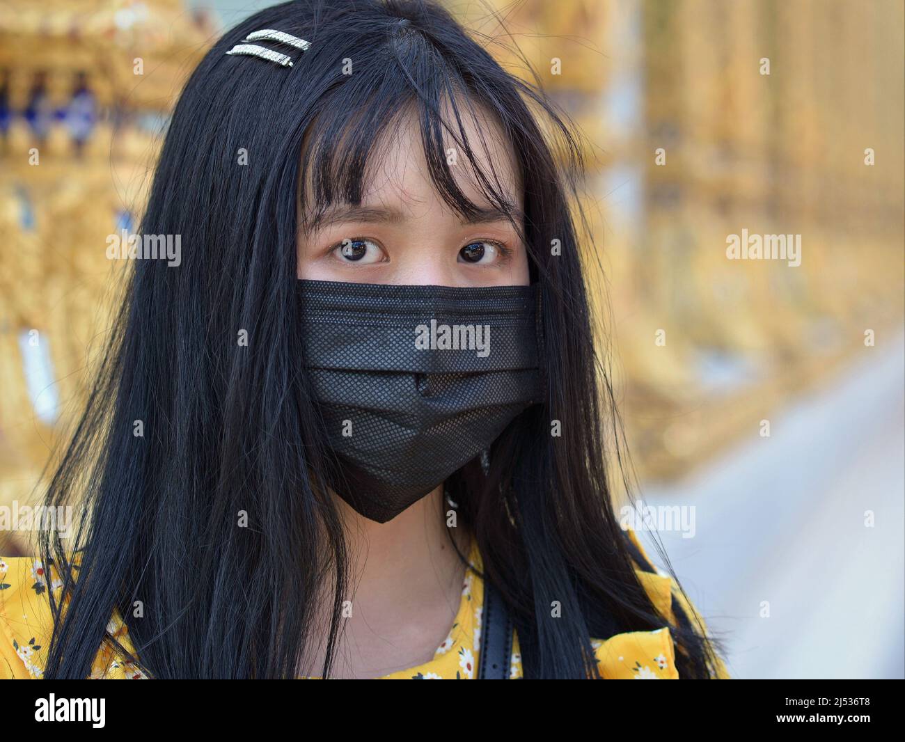 Attractive Thai girl with beautiful dark eyes and long black hair wears a black disposable protective face mask in front of a Buddhist wat temple. Stock Photo