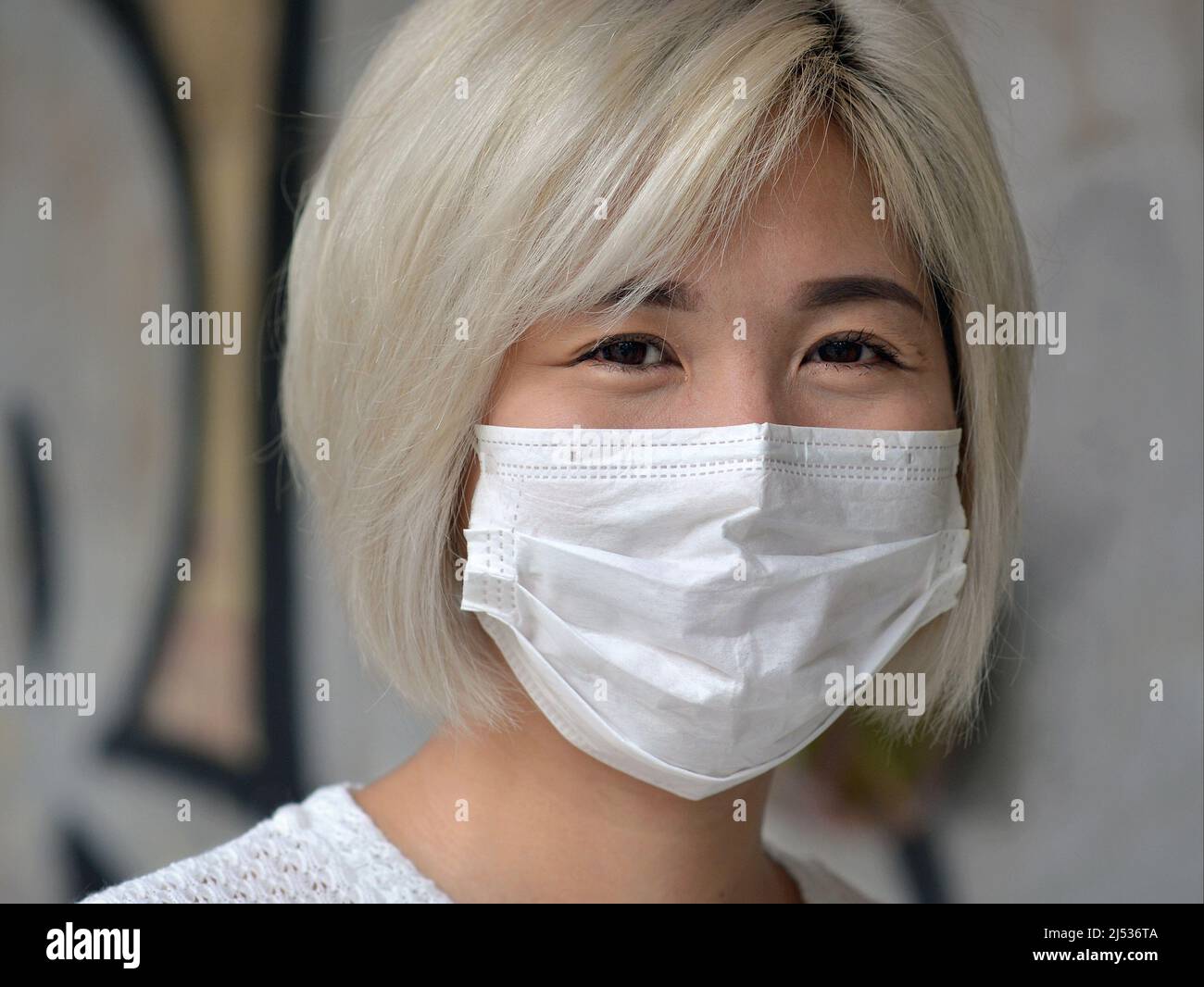 Beautiful young Vietnamese woman with silver white dyed hair and smiling eyes wears a disposable surgical face mask during the coronavirus pandemic. Stock Photo