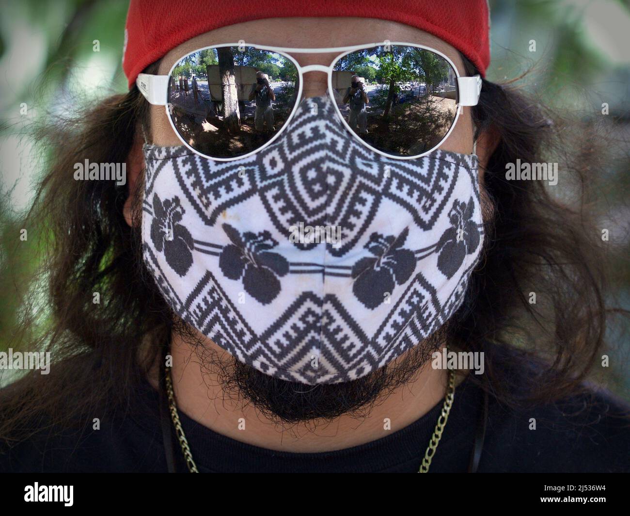 Young Caucasian man with long hair covers his face with non-medical cloth face mask and wears mirrored drop-shaped aviator sunglasses during pandemic. Stock Photo