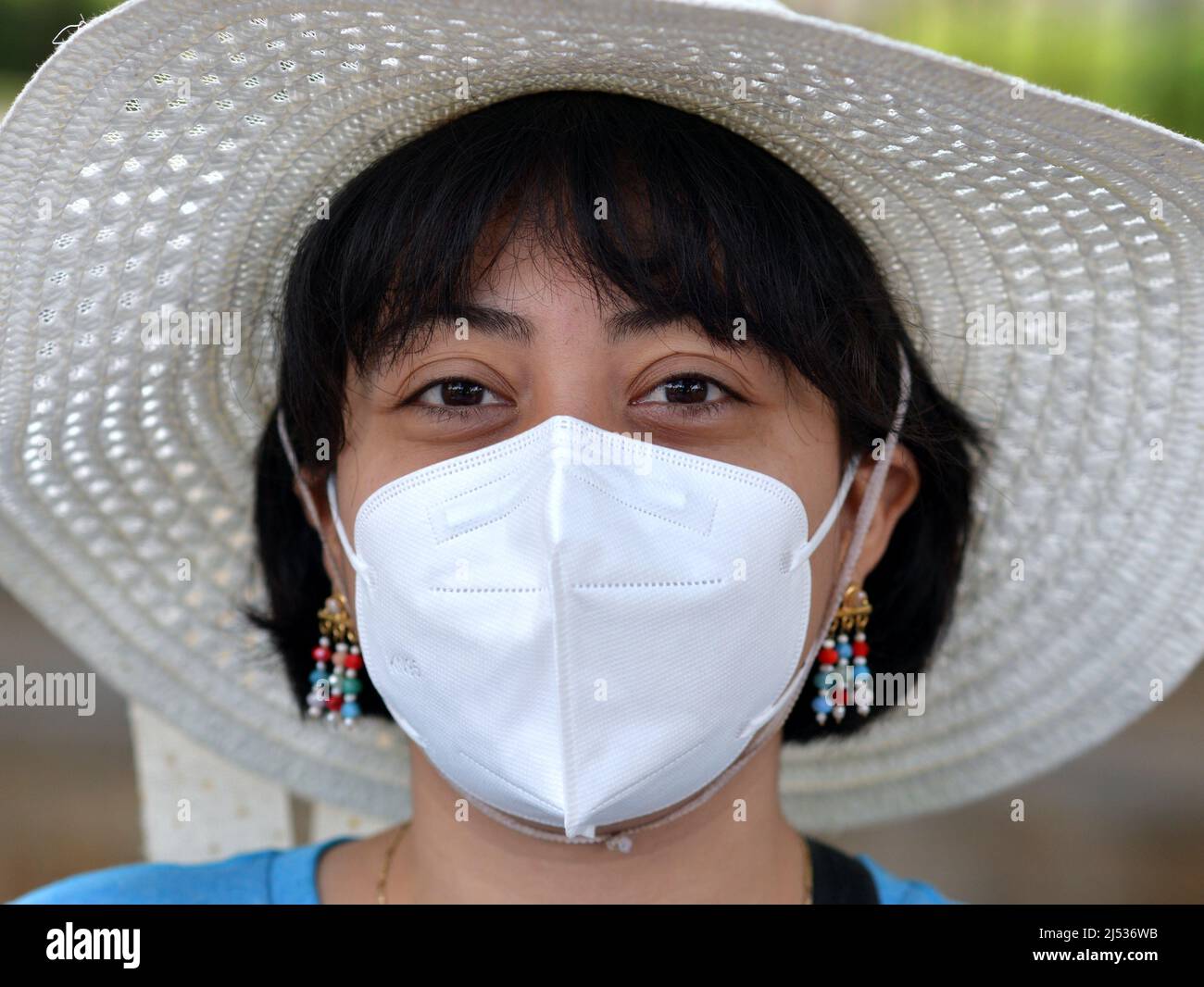 Young pretty Mexican woman with bob haircut, earrings and brown eyes wears white sun hat and white KN 95 face mask during global coronavirus pandemic. Stock Photo