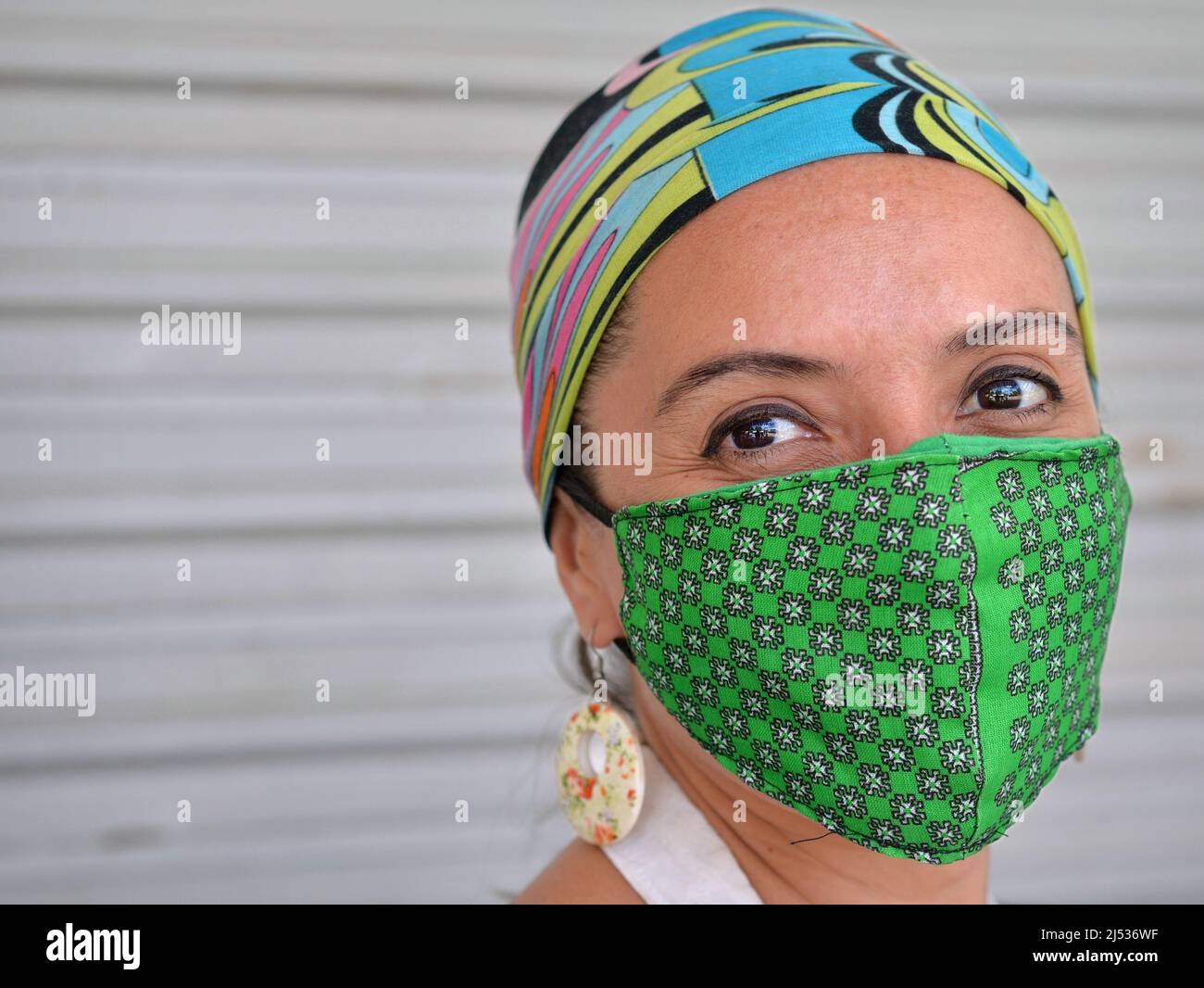 Young positive Caucasian woman with beautiful brown eyes wears a bandana and a green washable cloth face mask during the global coronavirus pandemic. Stock Photo