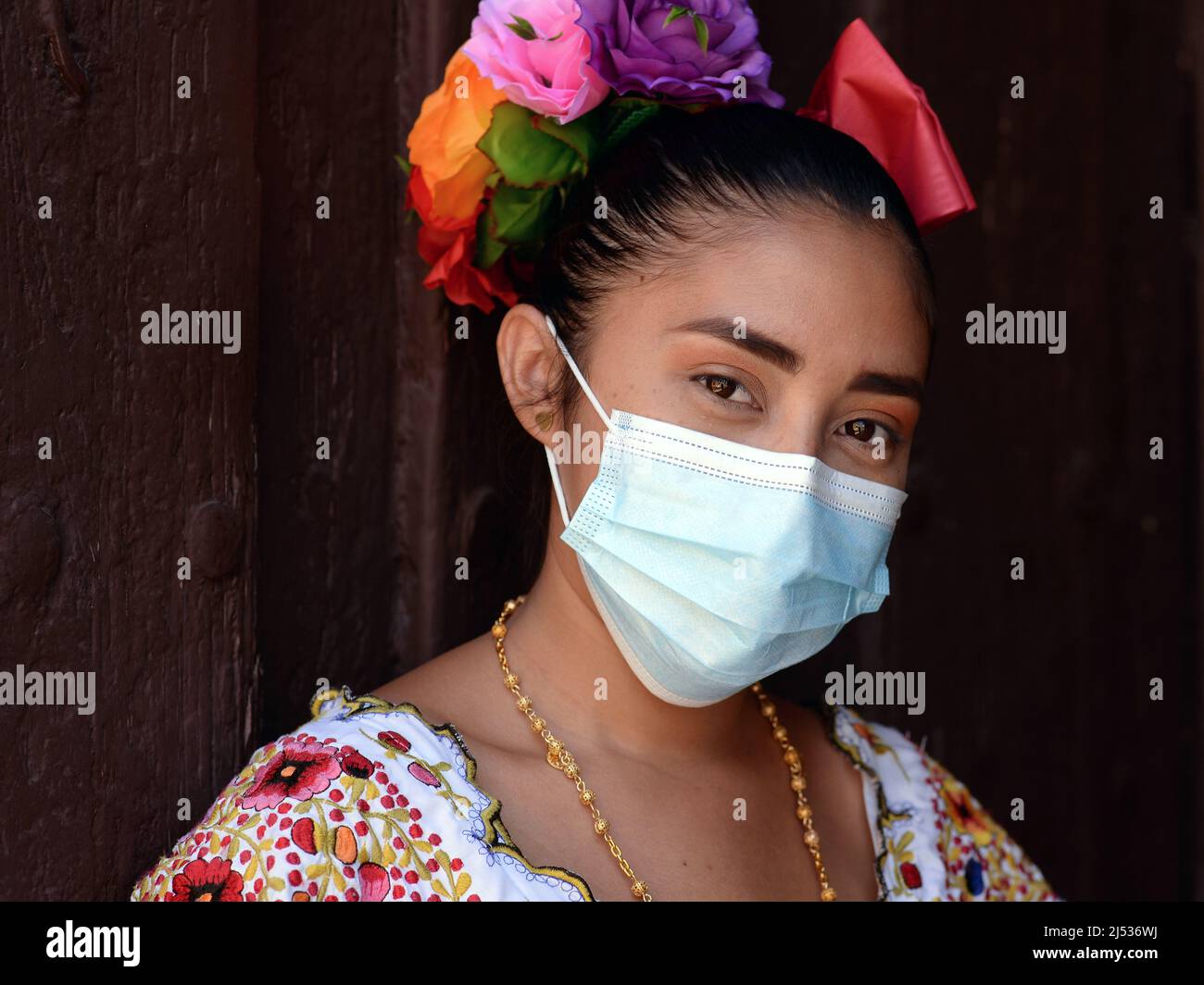 Attractive young Mexican woman with flowers in slicked-back hair wears a colorful traditional Yucatan Maya huipil dress and surgical face mask. Stock Photo
