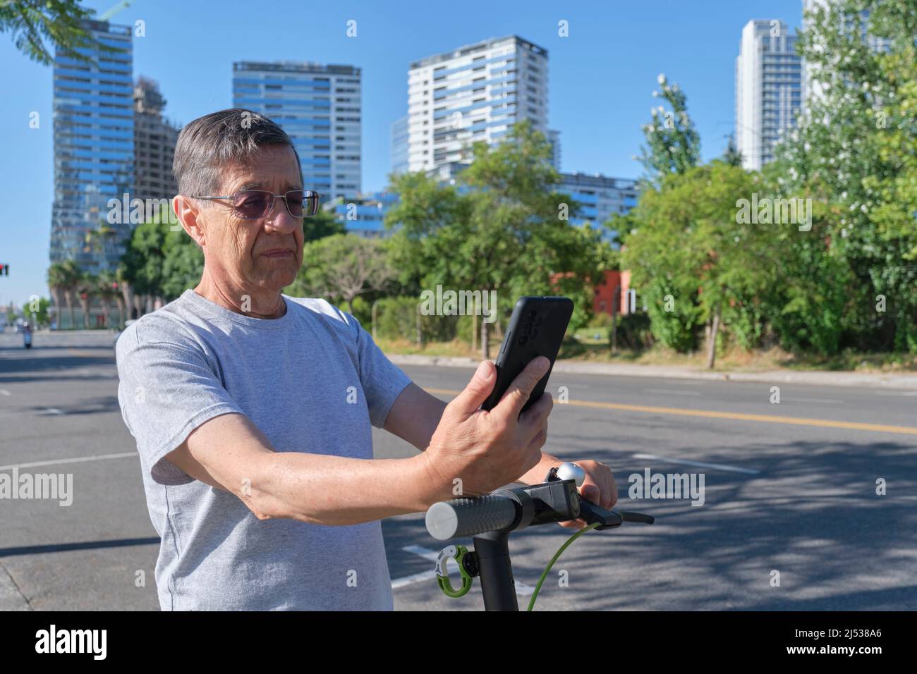 Hispanic senior man looking at his smart phone before riding his electric kick scooter in the city a sunny morning. Concepts of technology, active ret Stock Photo