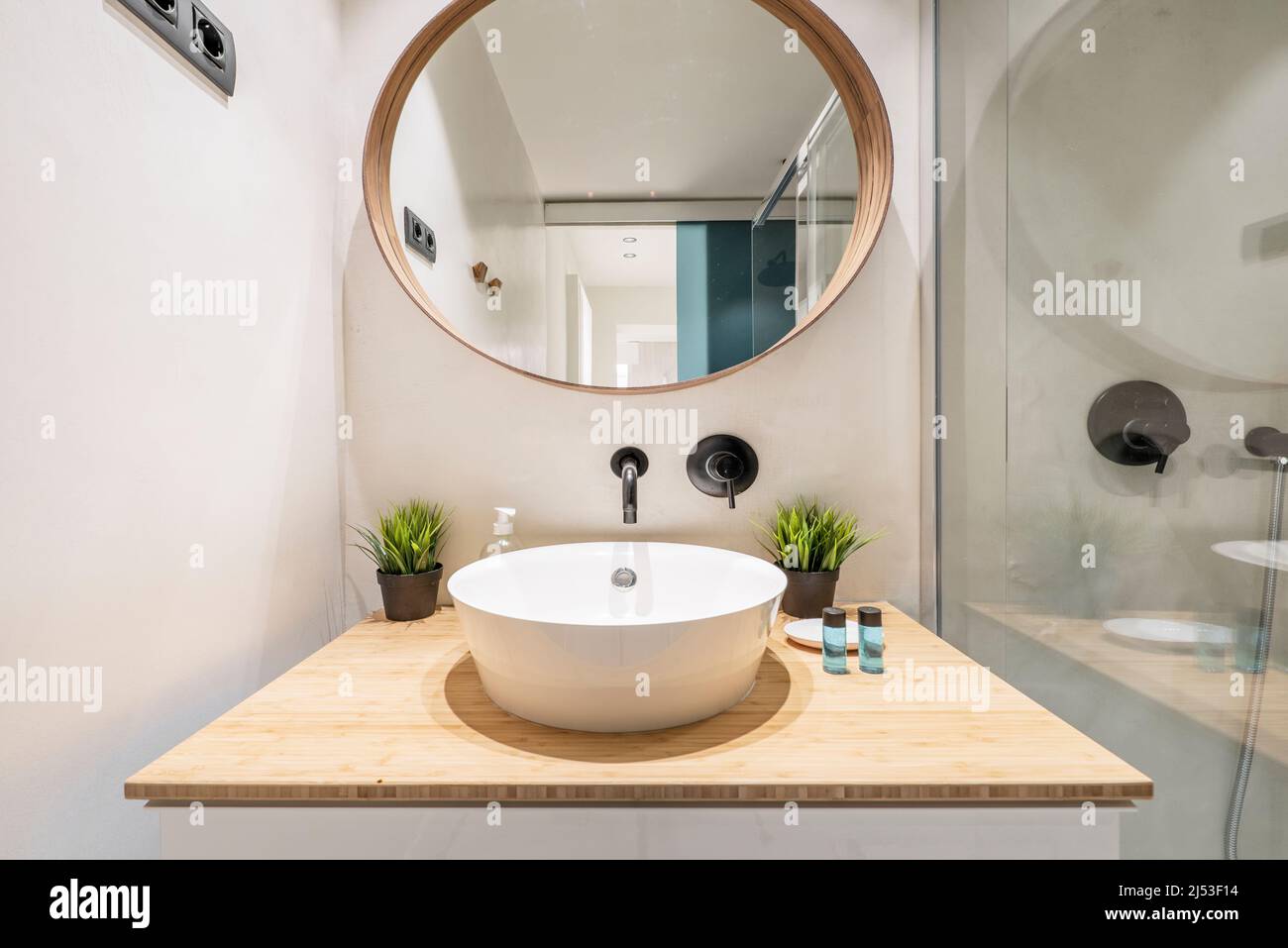 Bathroom with glossy wooden wall unit, light varnished wooden countertop, decorative plants and circular mirror with wooden frame and shower cabin wit Stock Photo