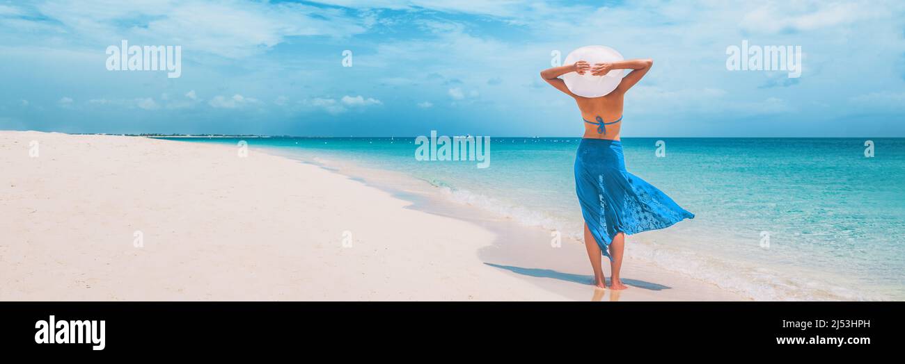 Beach vacation happy woman walking on summer travel Caribbean holiday with arms behind head wearing sun hat and sarong skirt. Ocean panoramic banner Stock Photo