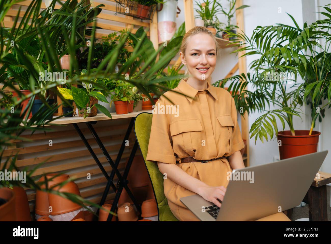 Portrait of attractive young businesswoman floral store owner sitting with laptop among green plants, looking at camera. Stock Photo