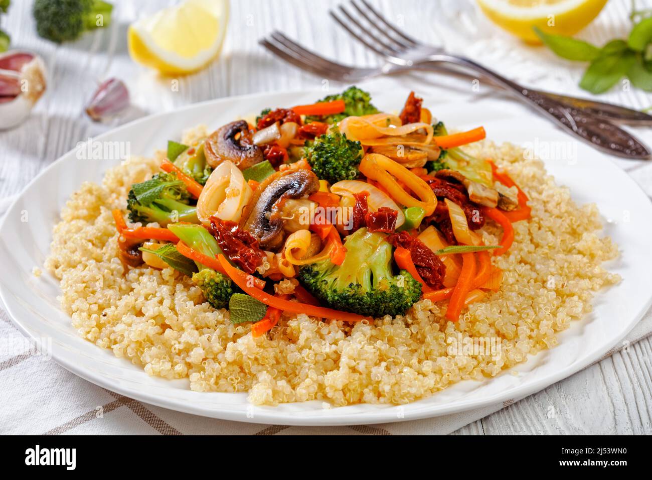 quinoa topped with stir fried broccoli, julienne carrots, sun dried tomatoes, leek and mushrooms on white plate on white wooden table with ingredients Stock Photo