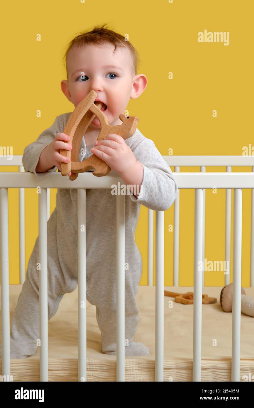 Sad toddler baby boy in the crib gnawing a wooden toy, yellow studio background. A tired child in pajamas Stock Photo