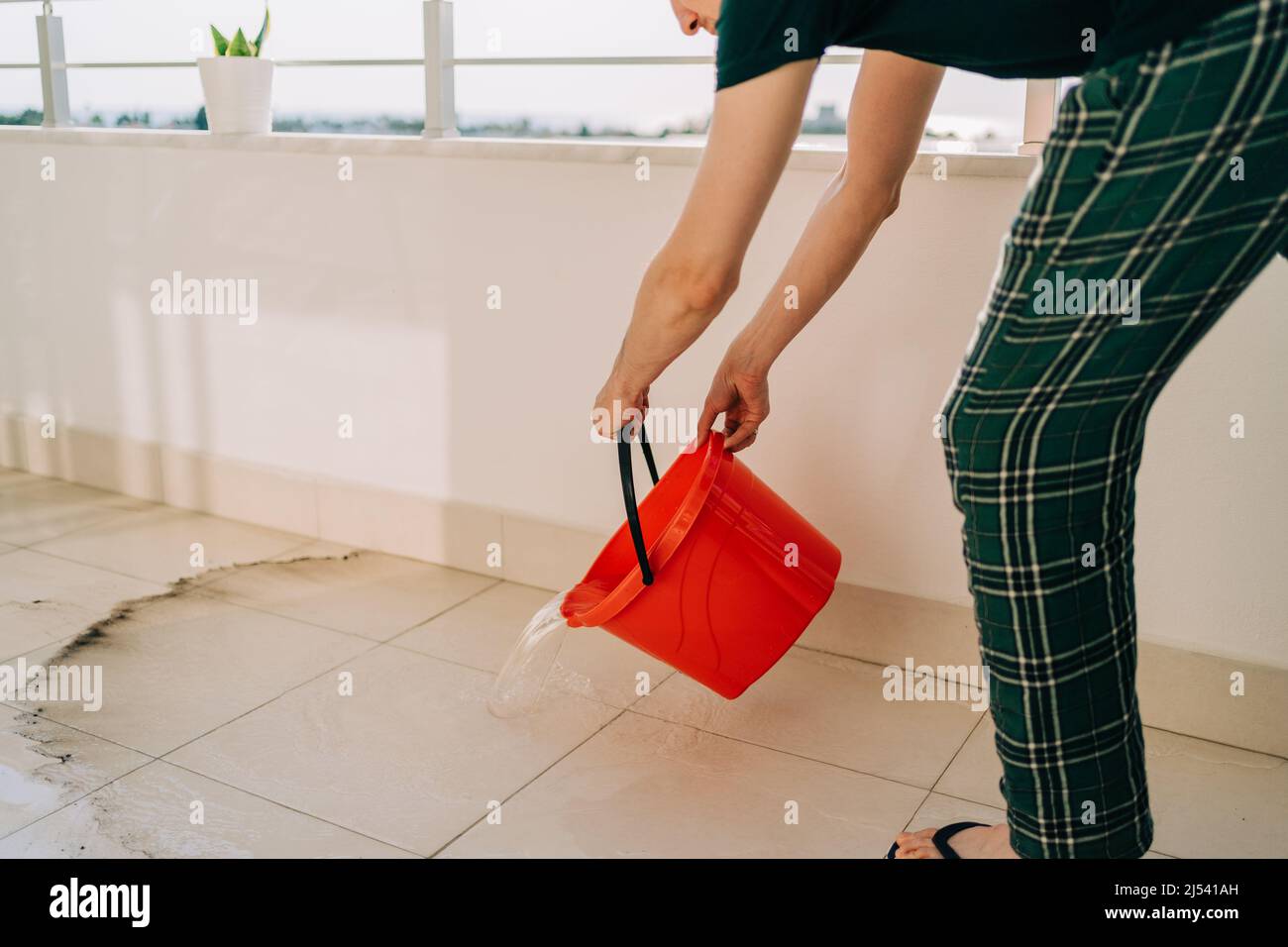 Man spilling the water from red bucket onto the tiled floor on the balcony in sunny day. Adult male doing home chores washing floors on the patio Stock Photo