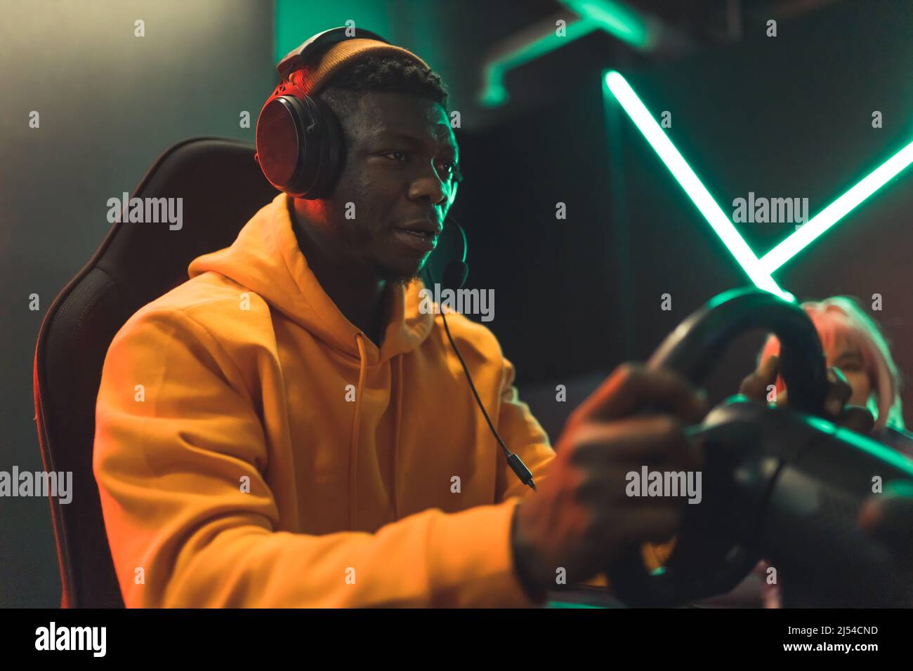 Adult African-American man in yellow hoodie and big black headphones racing on car simulator in gaming cafe. High quality photo Stock Photo