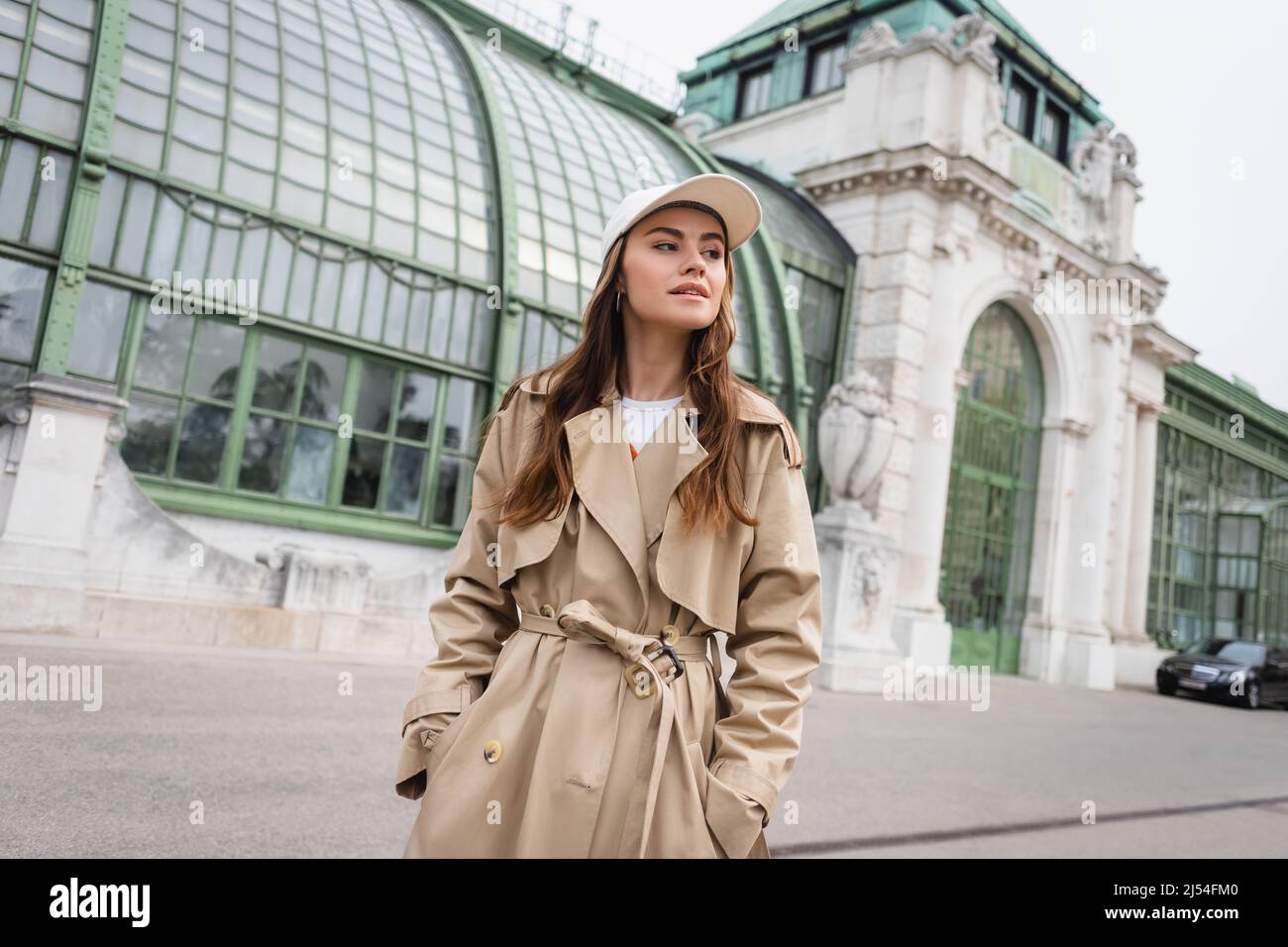 young woman in trench coat and baseball cap standing with hands in ...
