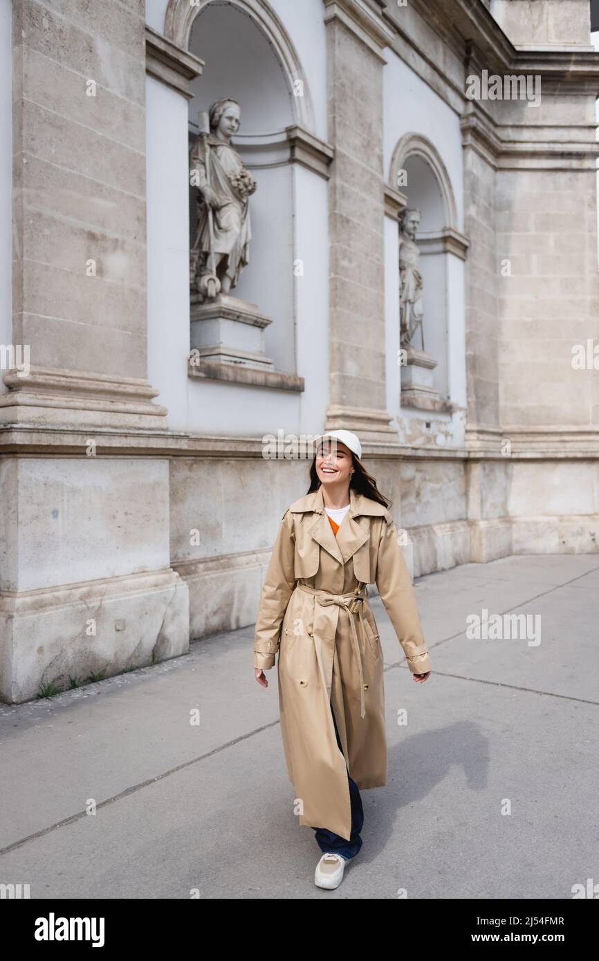full length of happy young woman in stylish trench coat and baseball