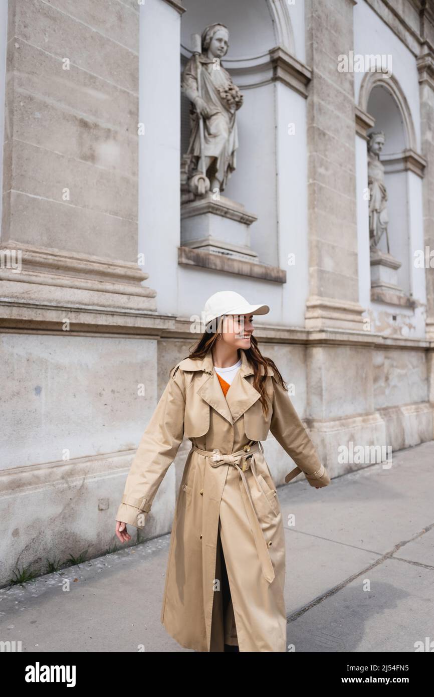 joyful young woman in stylish trench coat and baseball cap walking on
