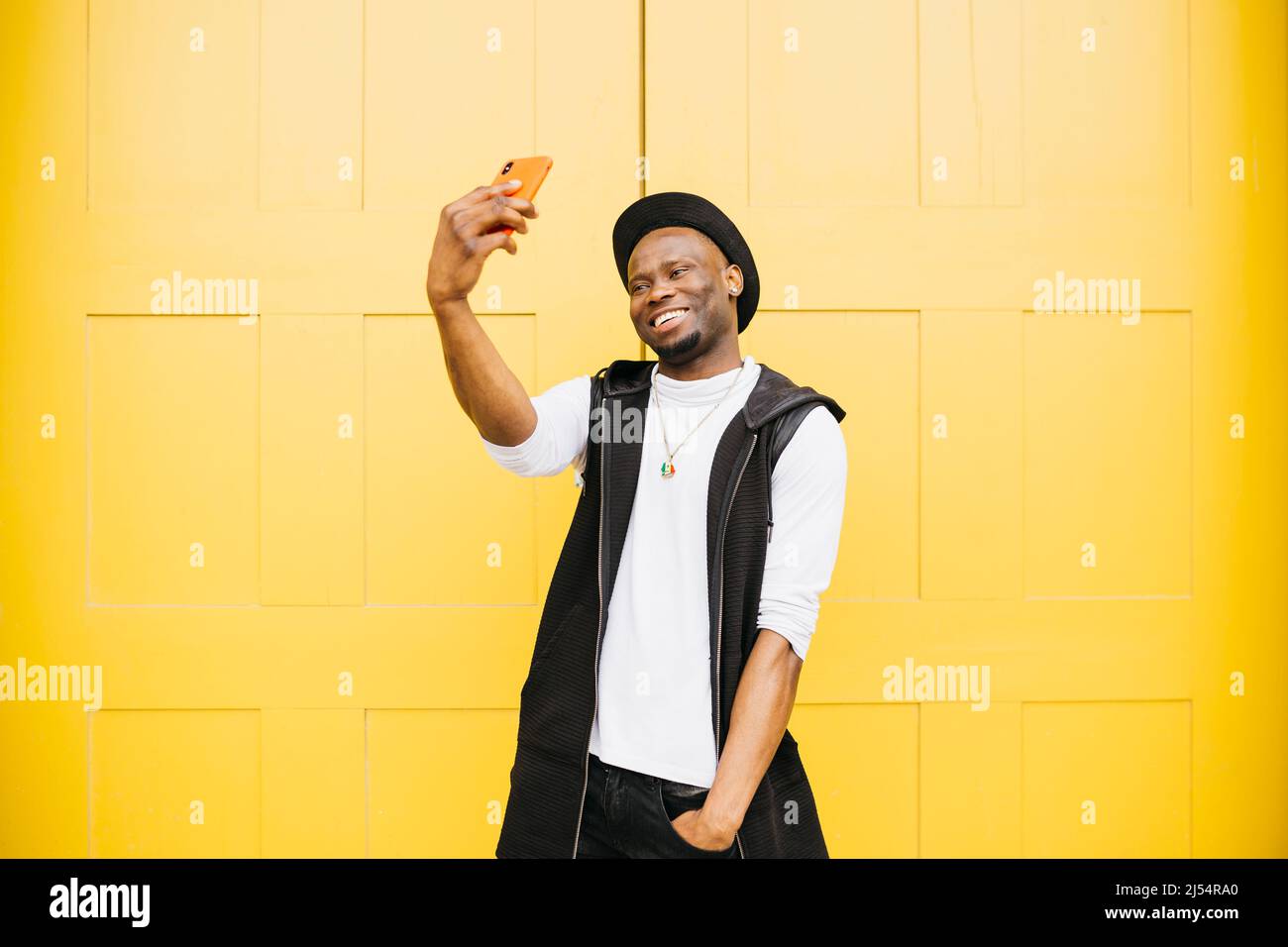 Portrait of a young smiling black male taking a selfie standing against a yellow door Stock Photo