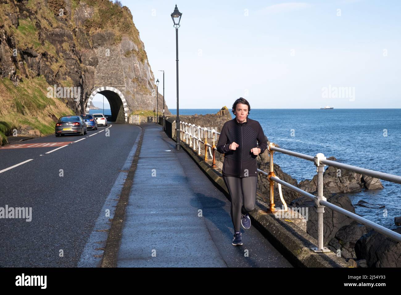 Woman dressed in dark clothing running alongside the ocean just outside Drains Bay on the Larne Coast in County Antrim. Northern Ireland. Stock Photo