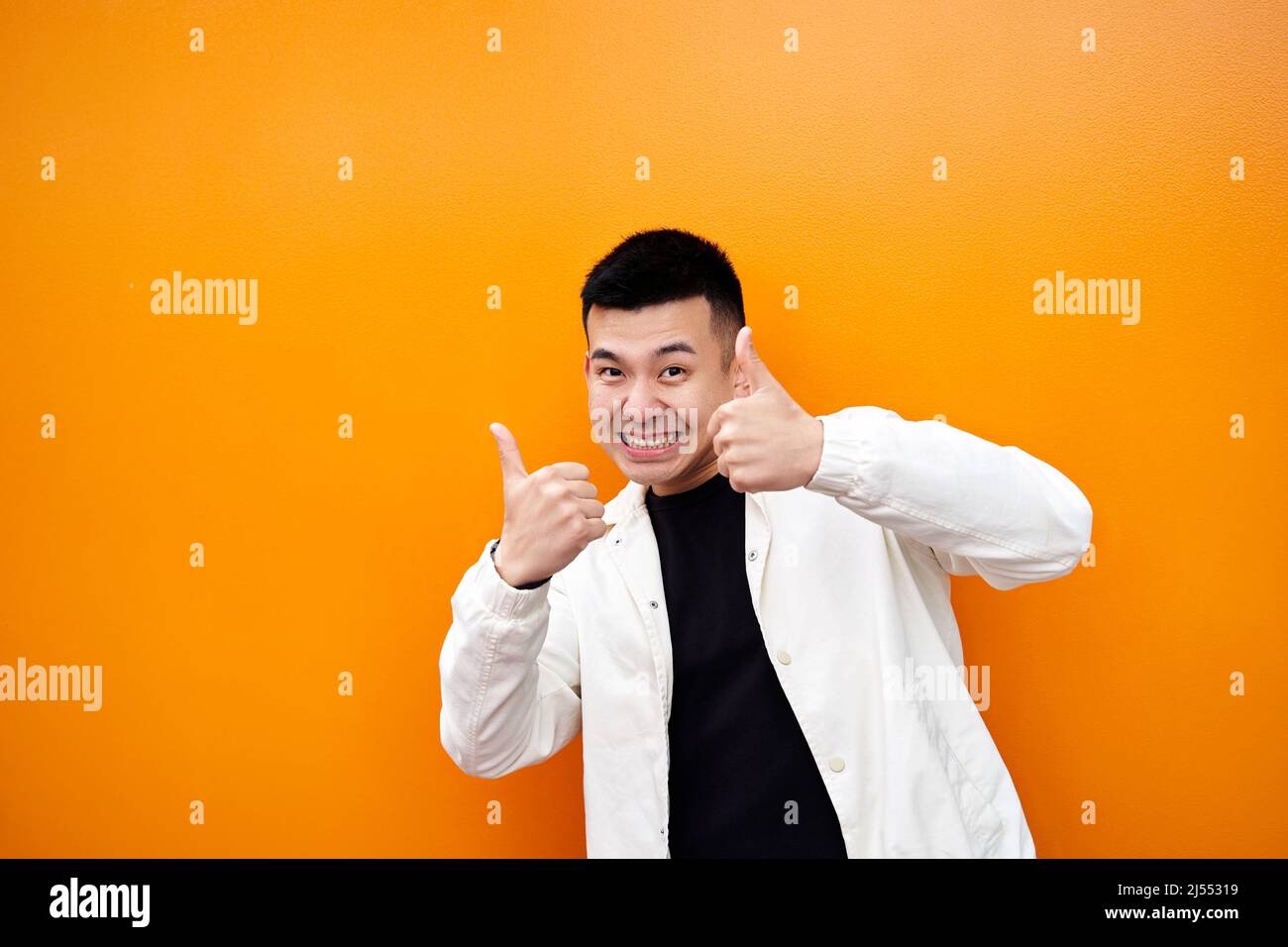 Good job man, I like it. Happy young Chinese man showing thumbs up and smiling broadly, giving positive feedback, sharing his positive opinion about the orange wall. High quality photo Stock Photo