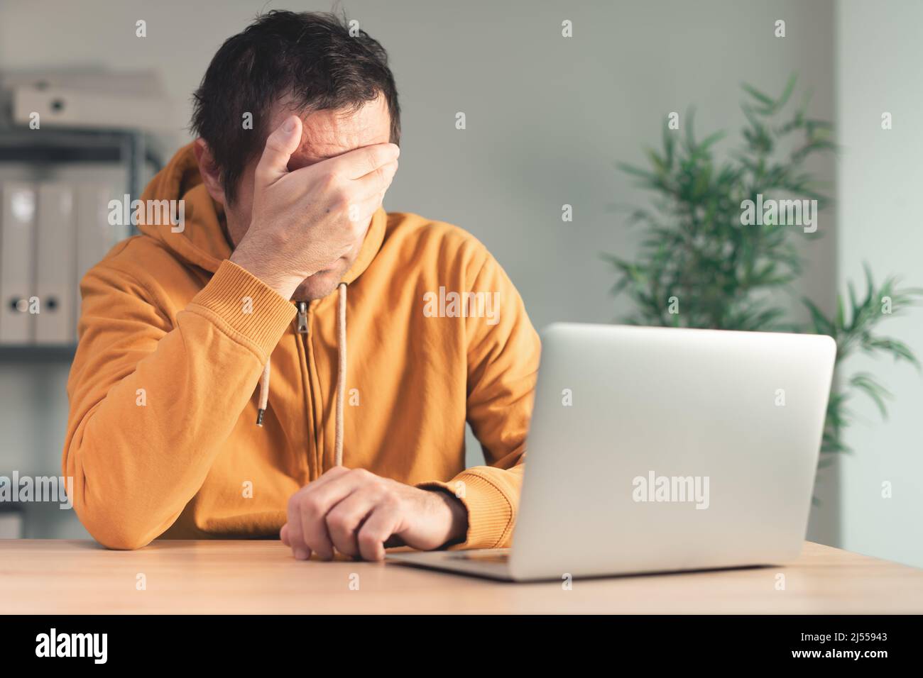 Disappointed freelancer at home office desk covering face with hand in front of laptop computer. Job problems and professional hardship concept. Stock Photo