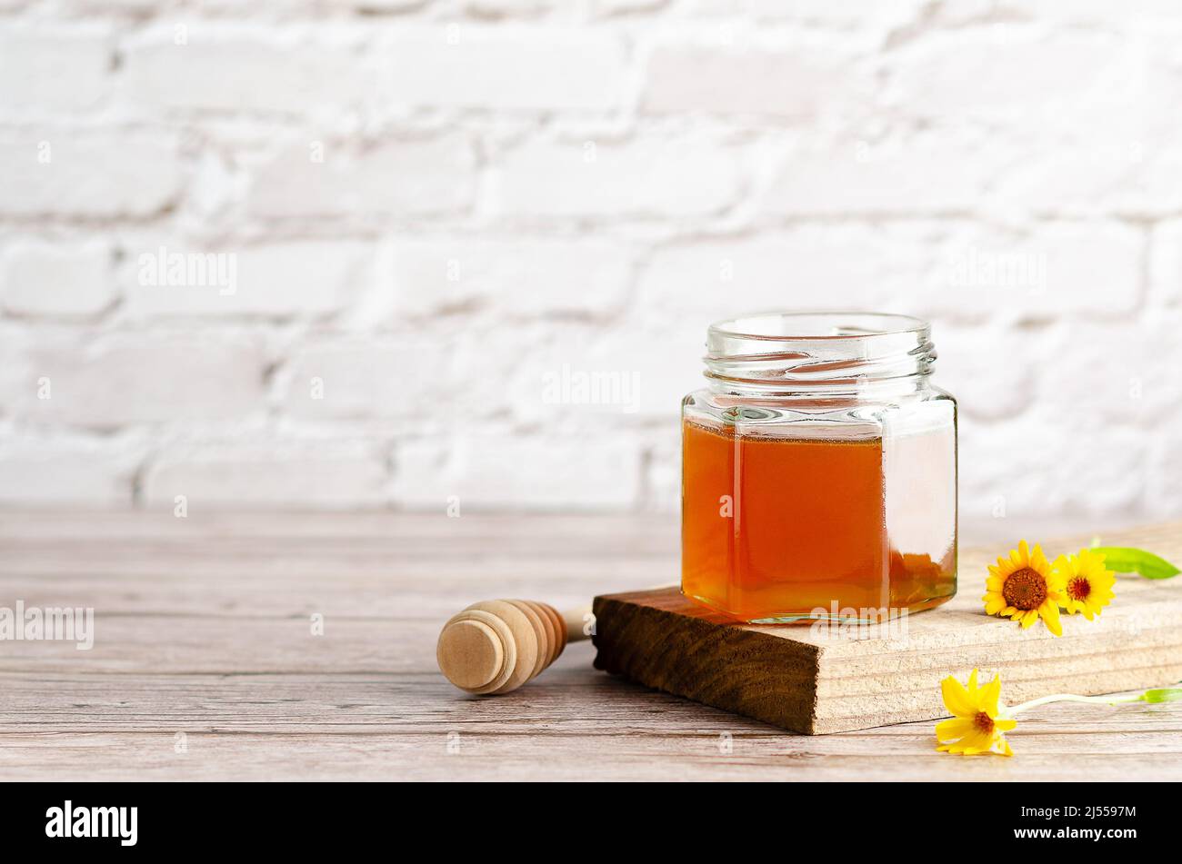 Honey in a jar on a piece of wood, a honey dipper and some marigolds, on a wooden table and a white wall with copy space. Stock Photo