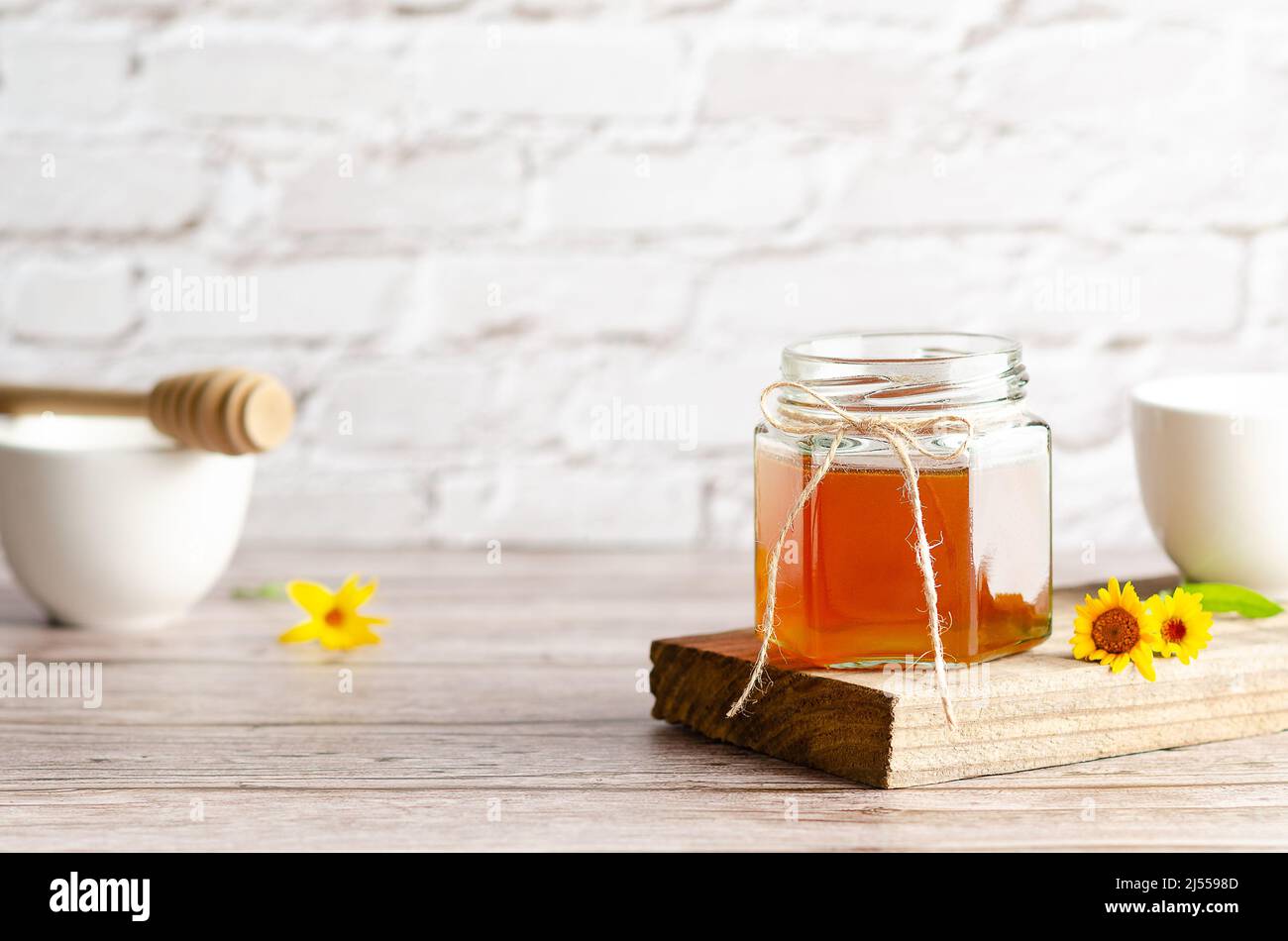 Honey in a jar on a piece of wood, a honey dipper, white bowls and some marigolds, on a wooden table and a white wall. Stock Photo