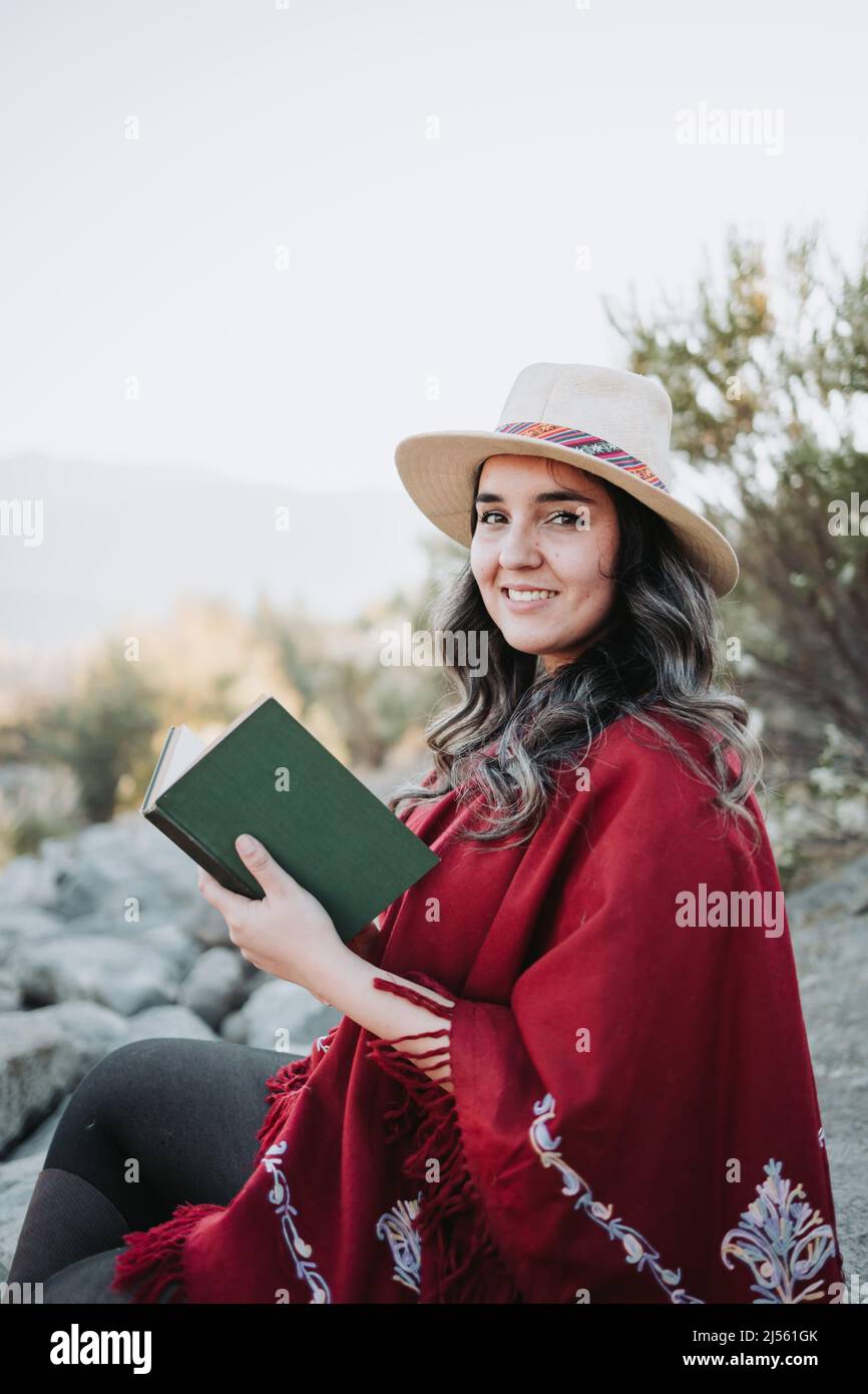 Young traditional hispanic woman using a hat and a red poncho, sitting in a natural space and reading a book. Stock Photo