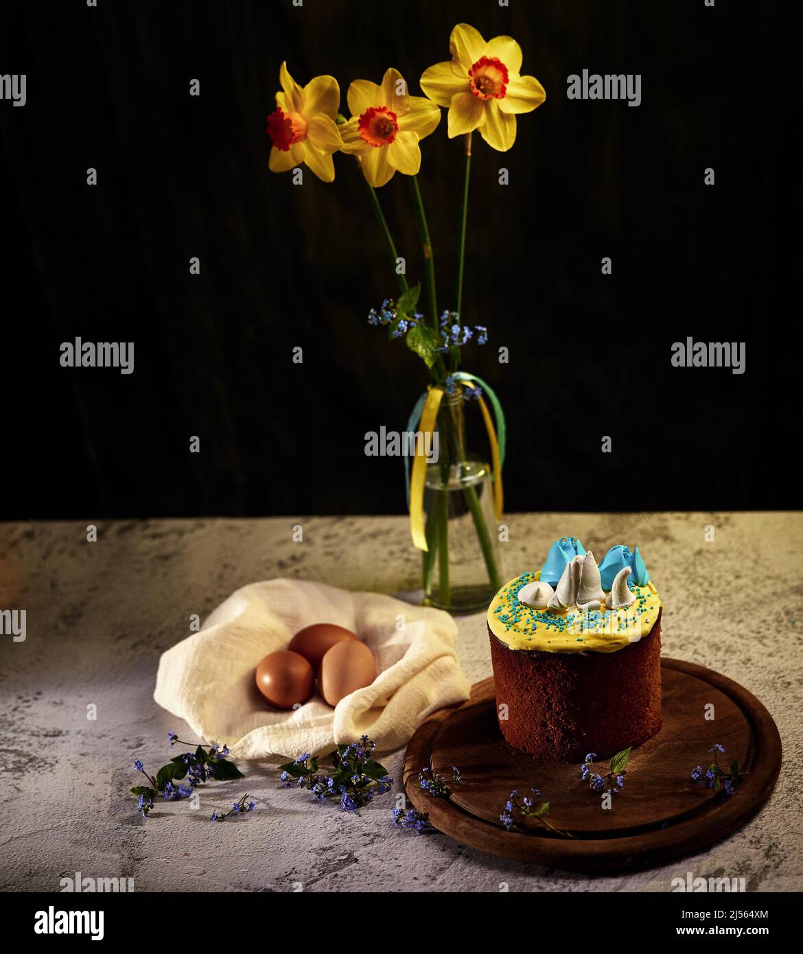 Easter board. Easter cake decorated with cream in the colors of the Ukrainian flag with blue flowers on a wooden board with eggs and daffodils in a va Stock Photo