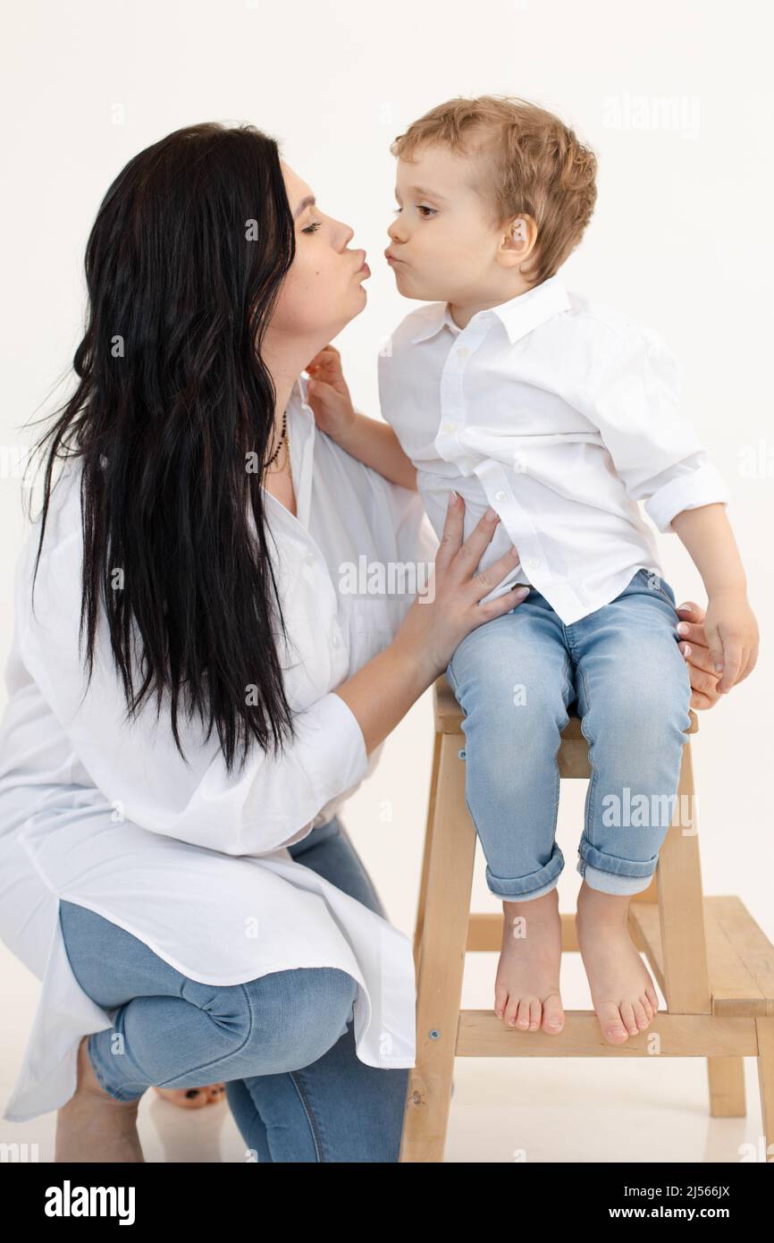 Vertical photo of dark-haired mom and barefoot son sending aerial kisses to each other. Boy standing on wooden chair Stock Photo