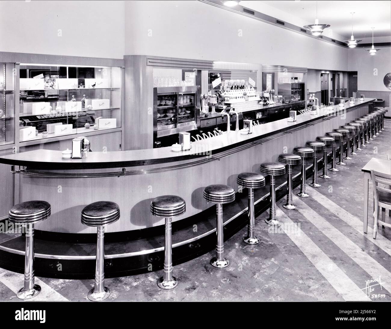 Soda shop, malt shop, soda fountain, lunch counter. A 1950s, cleanly-designed, long counter with about 20 rotating stools for seating. This shop doesn't have the really fancy art deco design of earlier years, but is lighter and brighter. The first soda fountain counters were created in the late 1800s, in pharmacies. The pharmacist served his customers mixed syrups and flavors mixed into chilled soda water, sometimes as a healthy drink and sometimes simply as a refreshing drink. Stock Photo