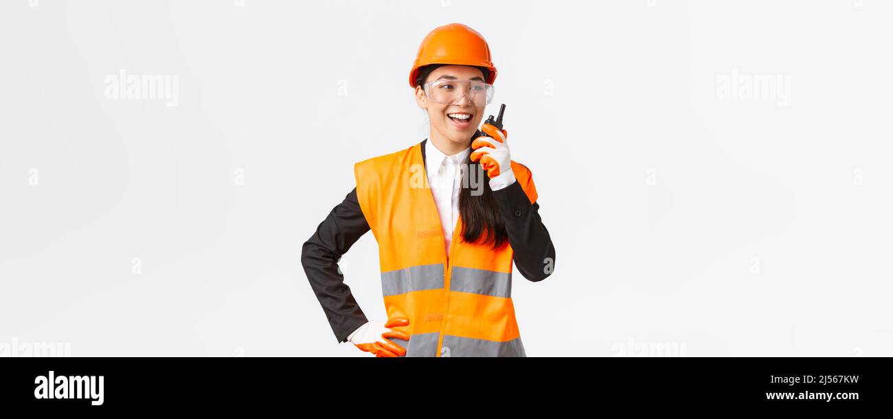 Confident pleased asian female engineer in safety helmet and uniform talking with chief architect using walkie-talkie. Satisfied construction Stock Photo