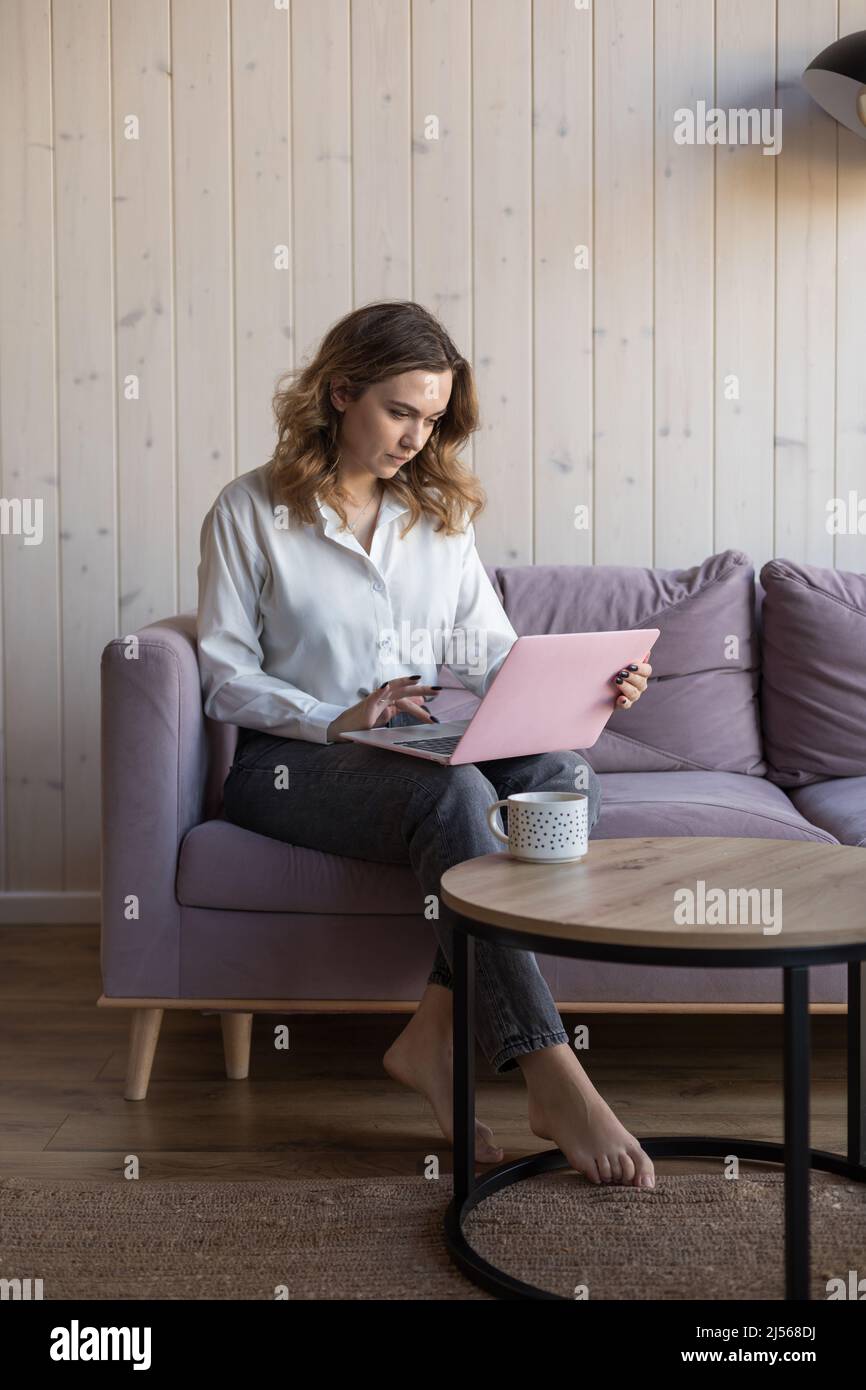 Portrait of woman working on modern pink laptop sitting on soft violet sofa in casual clothes near table with cup of coffee. Living room in elegant Stock Photo