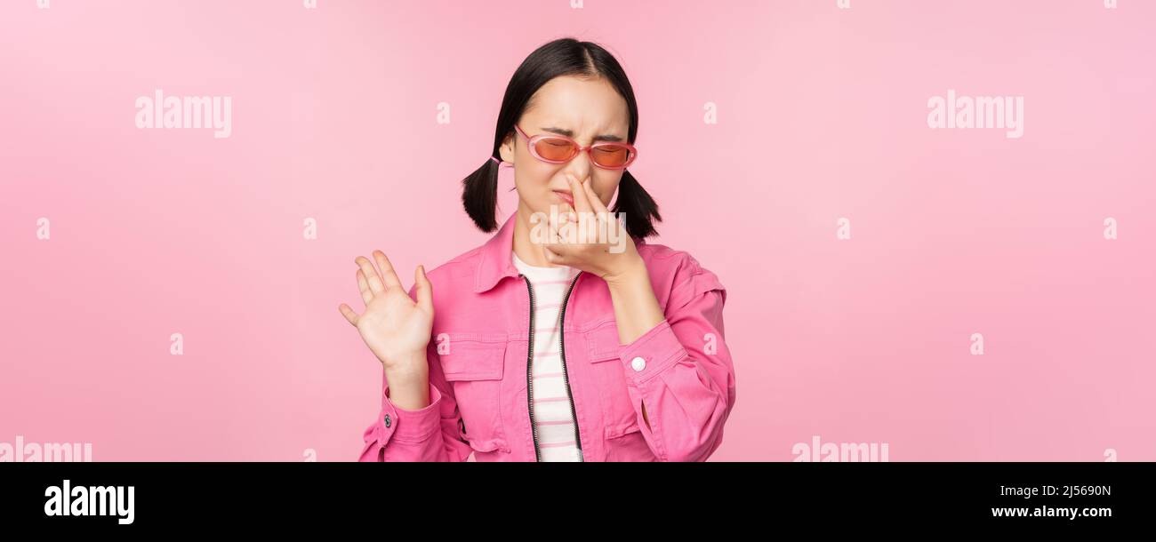 Stinky. Asian girl in sunglasses and stylish outfit, shuts her nose from disgust, dislike bad smell, standing over pink background Stock Photo